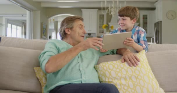 一个高个子白人男子坐在客厅的沙发上 转身和他年幼的孙子说话 孙子正站在沙发后面 给爷爷看他们都在看的平板电脑 动作缓慢 — 图库视频影像