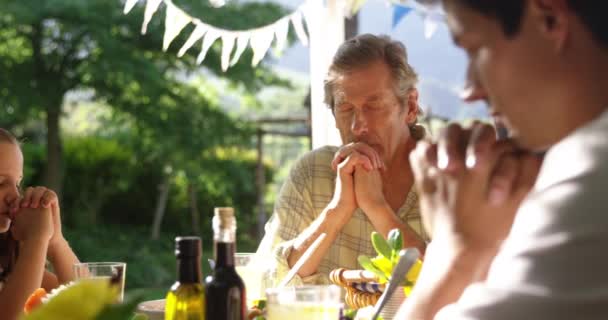 一个高个子白人男子和他的成年儿子 妻子和孙女坐在外面的餐桌前吃饭 两眼紧闭 双手紧握 饭前祈祷 动作缓慢 — 图库视频影像