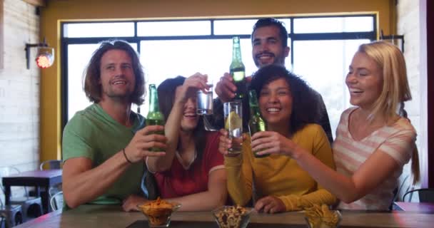 日中はパブのバーで多民族の男性と女性の友人5人のグループのフロントビュー スポーツゲームを見て ビールのボトルを保持し トーストを作るためにそれらを上げる スローモーションで一緒に笑顔とお祝いで笑って — ストック動画