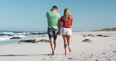 Sahilde birlikte boş vakit geçirmeyi seven mutlu beyaz bir çiftin arka planda mavi gökyüzü ve denizle el ele tutuşup ağır çekimde dolaşması.