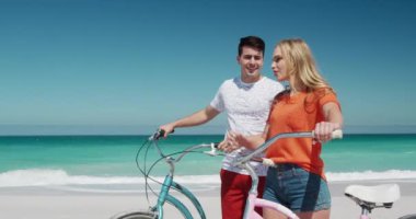 Kafkasyalı mutlu bir çiftin kumsalda boş vakit geçirmesinin yan görüntüsü. Bisikletleriyle yürüyor, mavi gökyüzü ve deniz ile arka planda ağır çekimde konuşup işaret ediyorlar.