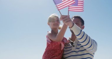Kafkasyalı mutlu bir adam ve kızının kumsalda birlikte boş vakit geçirmesinin ön görüntüsü, Amerikan bayrakları taşıyan, kızı taşıyan, arka planda ağır çekimde deniz ile gülümseyen adam.