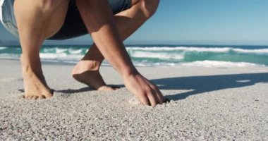 Arka planda mavi gökyüzü ve deniz ile sahilde çömelmiş, eli ağır çekimde kum toplayan beyaz bir adamın yan görüntüsü.