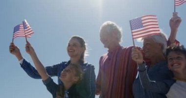 Sahilde birlikte boş vakit geçiren, arka planda mavi gökyüzü ile gülümseyen ve Amerikan bayraklarını tutan çok nesilli beyaz bir ailenin düşük açılı görüntüsü.