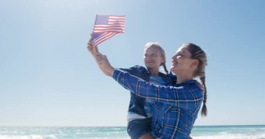 Kafkasyalı mutlu bir kadın ve kızı kumsalda birlikte boş vakit geçiriyorlar, ellerinde Amerikan bayrakları, kızı taşıyan bir kadın, arka planda mavi gökyüzü ve denizle gülümsüyor.