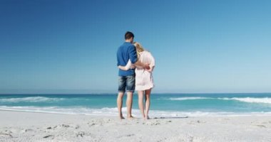 Sahilde birlikte vakit geçiren, ayakta duran, manzaraya hayran olan ve arka planda mavi gökyüzü ve denizle kucaklaşan mutlu, beyaz bir çiftin arka plan görüntüsü.