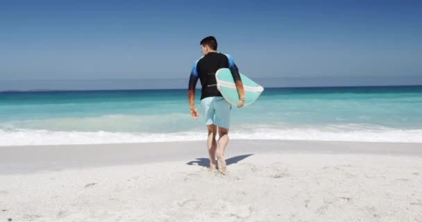 ビーチで自由な時間を楽しむ幸せな白人男性の背面ビュー スローモーションの背景に青い空と海と水の方に走るサーフボードを保持します — ストック動画