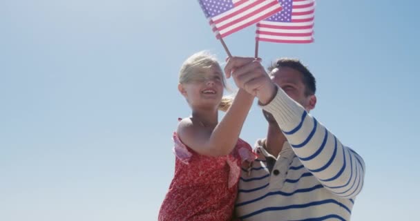 幸せな白人男性と彼の娘のフロントビュー一緒にビーチで自由な時間を楽しんで アメリカの国旗を持ち 女の子を運ぶ男 ゆっくりとした動きの中で海と笑顔 — ストック動画