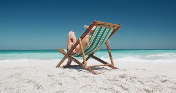 ビーチで自由な時間を楽しむ幸せな白人女性の背面ビュー 遅い動きの背景に青い空と海を背景にデッキチェアドリンクカクテルでビーチに座って — ストック動画