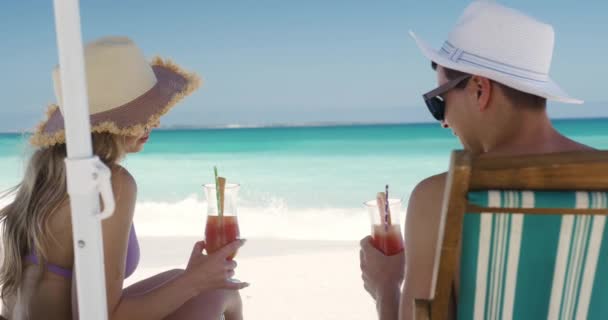 幸せな白人のカップルのサイドビュー一緒にビーチで無料の時間を楽しんで ゆっくりとした動きの背景に青い空と海を背景にパラソル飲料カクテルの下でデッキチェアに座って — ストック動画