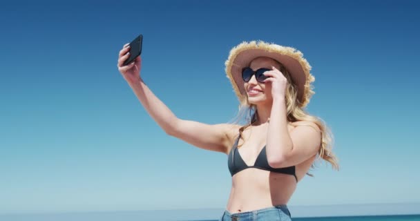 一个白人妇女在海滩上享受闲暇时光 戴着草帽和太阳镜站在海滩上 慢吞吞地拿着智能手机带着蓝天和大海自拍的低视角侧影 — 图库视频影像