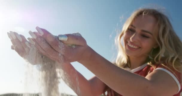 背景に青い空とビーチに身をかがめる幸せな白人女性の低い角度側のビュー 彼女の手で砂を選ぶ スローモーションで笑顔 — ストック動画