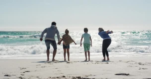 ビーチで一緒に自由な時間を楽しんでいる彼らの息子と娘と一緒に白人のカップルの後部ビュー ゆっくりとした動きの背景に海と青い空とジャンプし 水に直面して演奏 — ストック動画