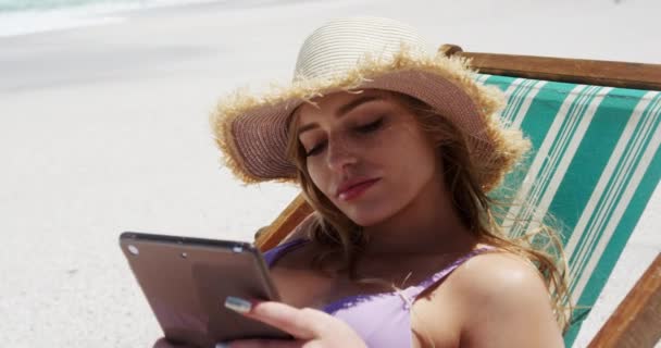 ビーチで自由な時間を楽しむ幸せな白人女性のフロントビューの終わり 遅い動きの背景に青い空と海とデジタルタブレットを保持デッキチェアでビーチに座って — ストック動画