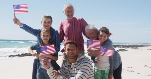 幸せな多世代の白人家族のフロントビュー一緒にビーチで自由な時間を楽しんで 立ち上がり 遅い動きの中で青空を背景に笑顔でアメリカの旗を保持 — ストック動画