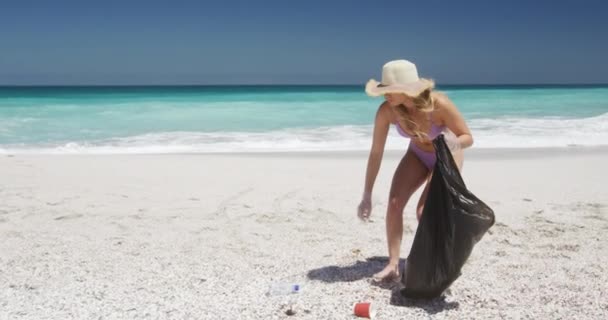 ビーチで自由な時間を楽しむ白人女性のフロントビュー ゴミを拾い スローモーションで背景に青い空と海とゴミ袋に入れて — ストック動画