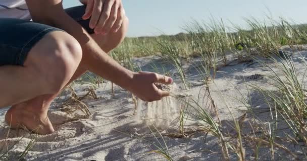 背景に青い空と海とビーチに身をかがめる白人男性の側のビューの低セクション スローモーションで彼の手で砂を選ぶ — ストック動画