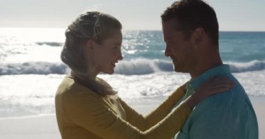 Sahilde birlikte vakit geçirmekten hoşlanan mutlu beyaz bir çiftin yan görüntüsü. Arka planda mavi gökyüzü ve deniz ile yavaş çekimde birbirlerine bakıyorlar.