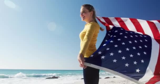 一个快乐的高加索女人在海滩上享受着闲暇时光 她站在那里 拿着美国国旗 慢吞吞地望着蓝天和大海 — 图库视频影像