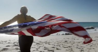 Sahilde boş vakit geçiren mutlu, beyaz bir kadının arka planda mavi gökyüzü ve denizle gülümseyen Amerikan bayrağını tutan suya doğru koşuşunun görüntüsü.