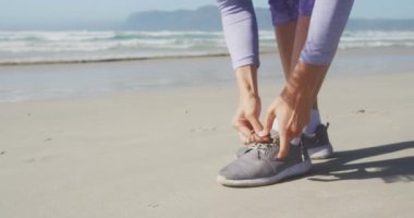 Güneşli bir günde deniz kenarında boş vakit geçiren, ayakkabı bağcıklarını yavaş çekimde bağlayan beyaz bir kadının ön tarafı.