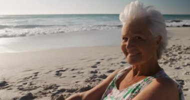 Güneşin altında kumsalda oturan, kameraya bakıp arka planda deniz ve mavi gökyüzü ile gülümseyen mutlu bir beyaz kadının portresi.