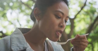 Düşük açılı kenar görüşlü, uzun siyah saçlı, şehir sokaklarında gezen, öğle yemeği yiyen, kot ceket giyen ve arka planda ağaçlarla birlikte ağaçlarla dolu bir caddede yürüyen melez bir kadının görüntüsü..
