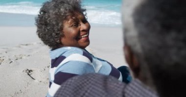 Arka planda mavi gökyüzü ve denizle sahilde oturan, birbirini kucaklayan, ağır çekimde gülümseyen yaşlı bir Afrikalı Amerikalı çiftin omzunun üstünden.