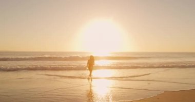 Gün batımında deniz kenarında sörf tahtasıyla koşan genç bir melez kadının ön görüntüsü.