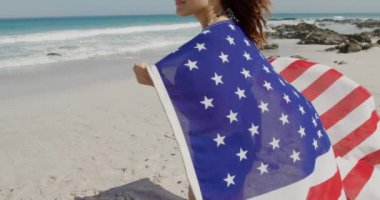 Deniz kenarında güneşli bir sahilde Amerikan bayrağıyla yürüyen genç bir kadının yan görüntüsü.