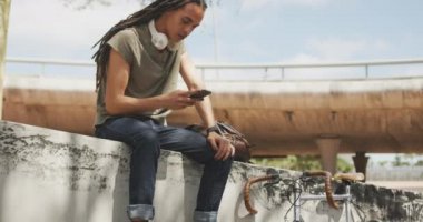 Güneşli bir günde, uzun saçlı ve şehirde gezen melez bir ırkın yan görüntüsü. Merdivenlerde oturuyor, gülümsüyor, akıllı telefon kullanıyor, bisikleti yavaş çekimde duvara dayanıyor..