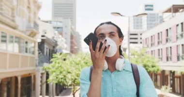 Uzun saçlı, güneşli bir günde şehirde gezen, Coronavirus maskesi takan ve akıllı telefonu yavaş çekimde kullanan melez bir ırkın ön görüntüsü..