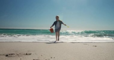 Güneşli bir sahilde koşan ve elinde bir kova kürek tutan beyaz bir kızın ön görüntüsü. Mavi gökyüzü ve deniz arka planda ağır çekimde.