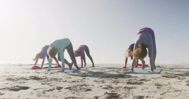 Güneşli bir günde kumsalda boş vakit geçiren bir grup beyaz kadın arkadaşın önünde diz çöküp ağır çekimde yoga çalışması yapıyorlar.