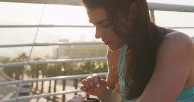 Uzun siyah saçlı, spor giyimli, kentte egzersiz yapan, kulaklıkla müzik dinleyen, köprüde oturup, saatini yavaş çekimde kontrol eden beyaz bir kadının yan görüntüsü..
