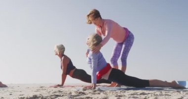 Güneşli bir günde deniz kenarında boş vakit geçiren bir grup beyaz kadın arkadaşın yan görüntüsü. Yoga yapıyorlar. Yoga yapan kadın yoga eğitmeni onlara ağır çekimde yardım ediyor.