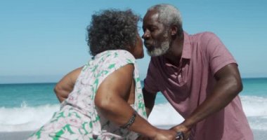 Arka planda mavi gökyüzü ve deniz ile sahilde durmuş yavaş çekimde öpüşen yaşlı Afrikalı Amerikalı çiftin yan görüntüsü.