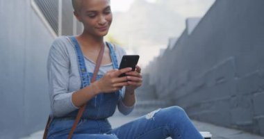 Güneşli bir günde şehirde kısa boyalı sarı saçlı mutlu melez bir kadın, akıllı telefonunu yavaş çekimde merdivenlerde oturuyor..