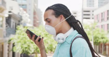 Uzun saçlı, güneşli bir günde şehirde gezen, Coronavirus maskesi takan ve akıllı telefonu yavaş çekimde kullanan melez bir ırkın yan görüntüsü..