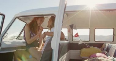 Deniz kenarında güneşli bir plajda, bir karavanda öpüşen beyaz bir kadın ve erkeğin kucağında oturan bir kadının yan görüntüsü.