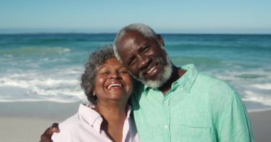 Arka planda mavi gökyüzü ve deniz ile sahilde dikilen, birbirlerine sarılan, kameraya bakan ve ağır çekimde gülümseyen yaşlı bir Afrikalı Amerikalı çiftin portresi.
