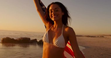 Genç bir melez kadının ön görüntüsü. Gün batımında deniz kenarında Amerikan bayrağıyla koşuyor.