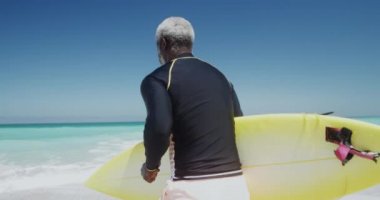 Yaşlı bir Afrikalı Amerikalı adamın güneşin altında bir plajda koşarken ve kolunun altında bir sörf tahtası taşırken, arka planda mavi gökyüzü ve deniz ağır çekimde.