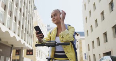 Güneşli bir günde kısa boyalı sarı saçlı, şehirde gezen, akıllı telefonunu kullanan, kulaklıkla müzik dinleyen, yavaş çekimde binaları olan elektrikli scooter 'a yaslanan mutlu melez kadının düşük açılı ön görüntüsü..
