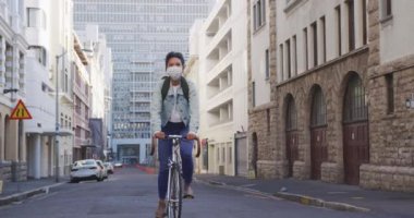 Uzun siyah saçlı, gün boyunca şehir sokaklarında gezen, hava kirliliği ve koronavirüse karşı maske takan, arka planda yavaş çekimde binalarla bisiklete binen melez bir kadının ön görüntüsü..