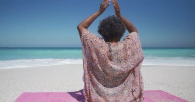 Kıdemli bir Afrikalı Amerikalı kadının, güneşin altında bir plajda oturup yoga yaparken, yoga pozu verirken, arka planda mavi gökyüzü ve deniz yavaş çekimde.
