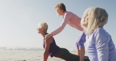 Güneşli bir günde deniz kenarında boş vakit geçiren bir grup beyaz kadın arkadaşın yan görüntüsü. Yoga yapıyorlar. Yoga yapan kadın yoga eğitmeni onlara ağır çekimde yardım ediyor.