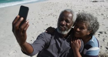 Sahilde oturan, arka planda deniz olan, kucaklayan ve gülümseyen yaşlı bir Afrikalı Amerikalı çiftin yüksek açılı yan görüntüsü. Adam ağır çekimde akıllı telefonla selfie çekiyor.