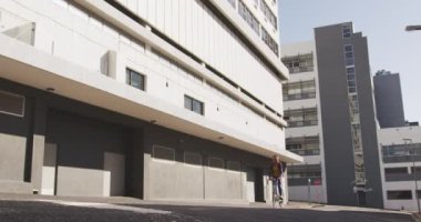 Düşük açılı, uzun siyah saçlı ve gündüzleri şehir sokaklarında gezen, arka planda yavaş çekimde binaları olan, bisikletine binen melez bir kadın..