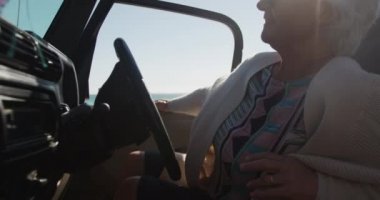 Yol gezisi sırasında plajda oturan, direksiyonda oturan, güneş gözlüğü takan, ağır çekimde gülümseyen yaşlı beyaz bir kadının yan görüntüsü.